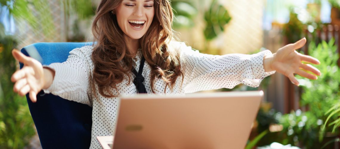 Green Home. smiling trendy middle aged housewife with long wavy hair in the modern living room in sunny day having video meeting on a laptop.